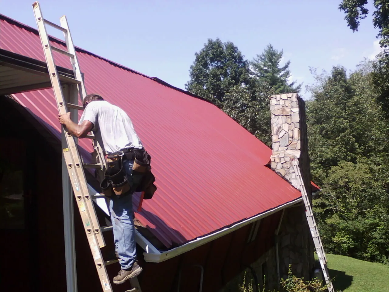 Roofer on a ladder beside a red metal roof and stone chimney — Soffit & Fascia Repair by Roofing Brothers in Gayville
